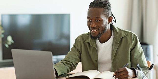 A young man is looking intently at his computer, with a pen in hand ready to jot notes in a notebook.
