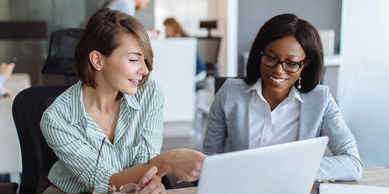 Two young women are pointing to and looking at a laptop. They are discussing what they see on the screen.