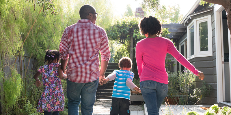 Family of Mom, Dad and two kids walking away from the camera towards their house. they are holding hands and are energetic.