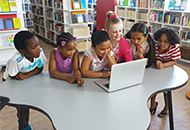 children in a school library gathered around a laptop on a table