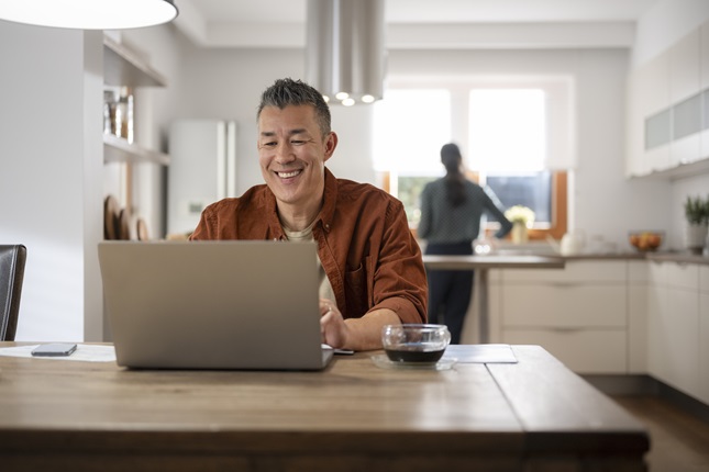 Man sitting at kitchen table, smiling at his open laptop.